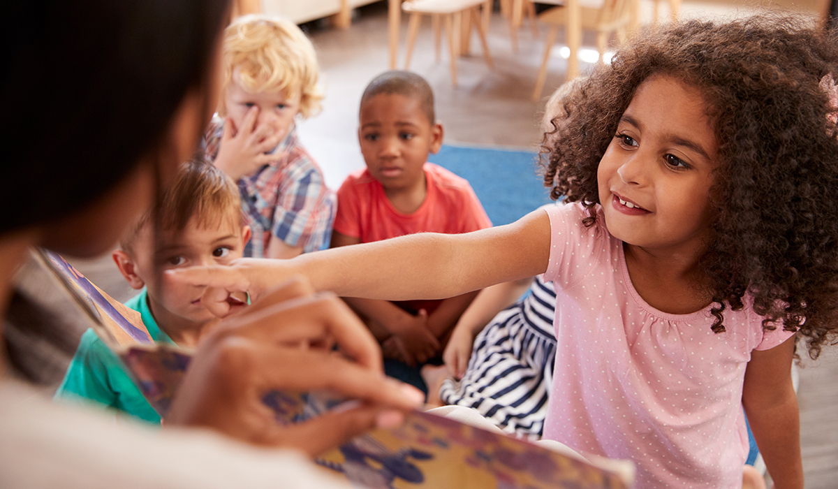 Toddler pointing at an object in a book her teacher is holding up. A group of kids sits behind her looking at the book