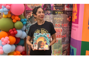 Author Xelena González holding her book Of the Sun in front of a pastel balloon display.