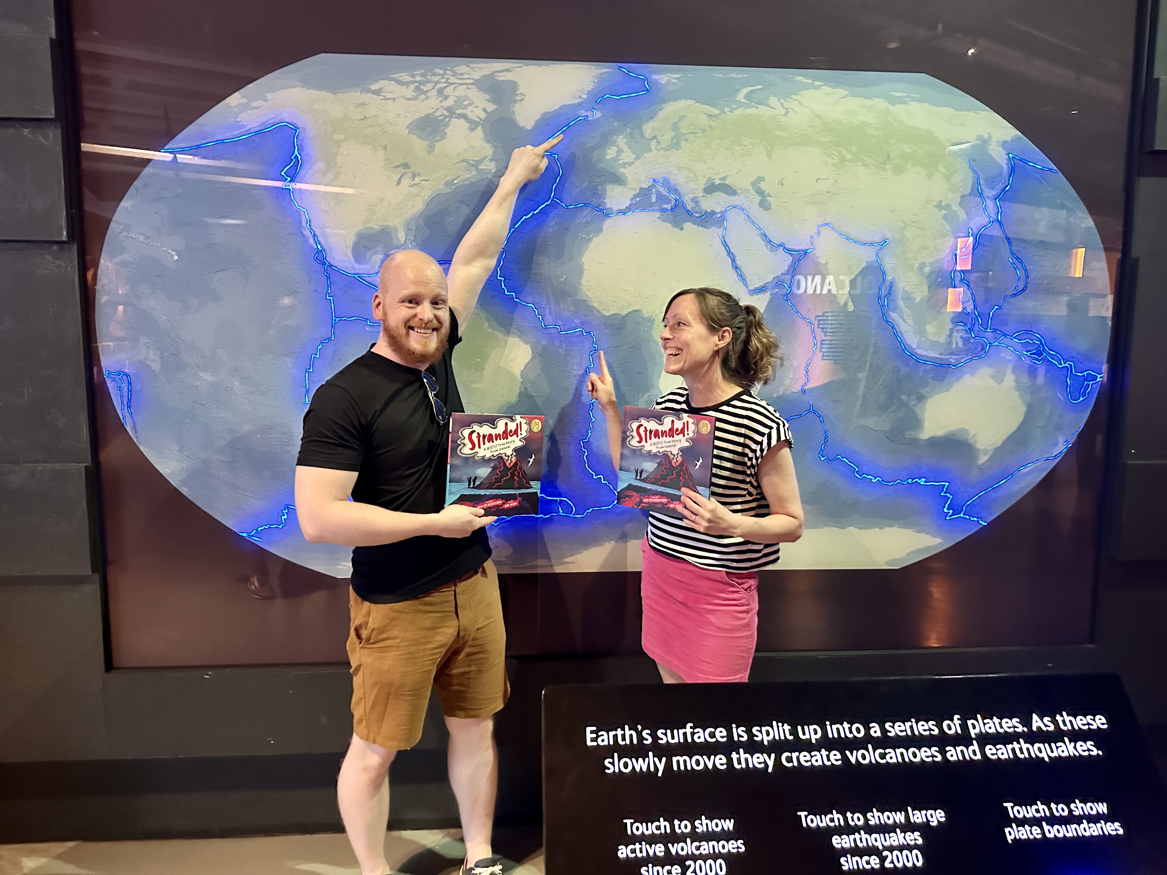 Icelandic Author Ævar Þór Benediktsson standing with his book, Stranded!, and pointing at Iceland on a map of the world