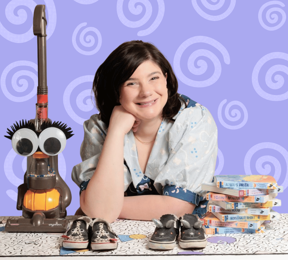 image of author Eleni Tassopoulos Wehner smiling at the camera and propping her chin up on a fist. A pile of books, two pairs of childrens shoes, and a vaccuume with large googly eyes lay on the table around her.