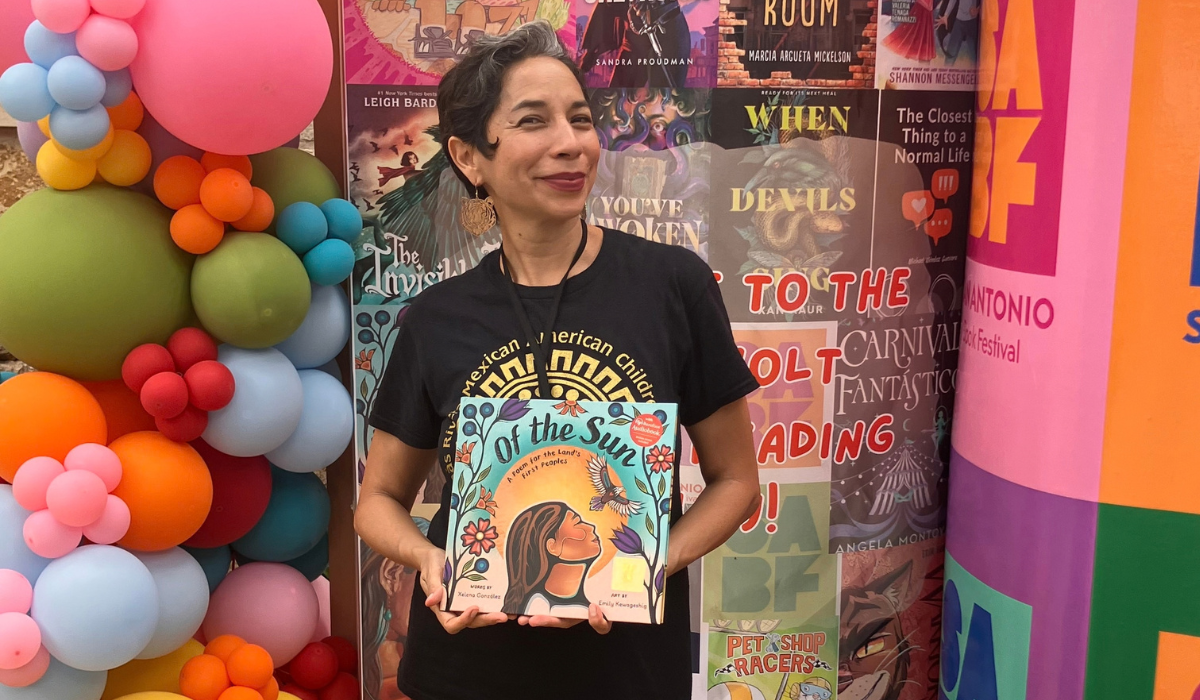 Author Xelena González holding her book Of the Sun in front of a pastel balloon display.